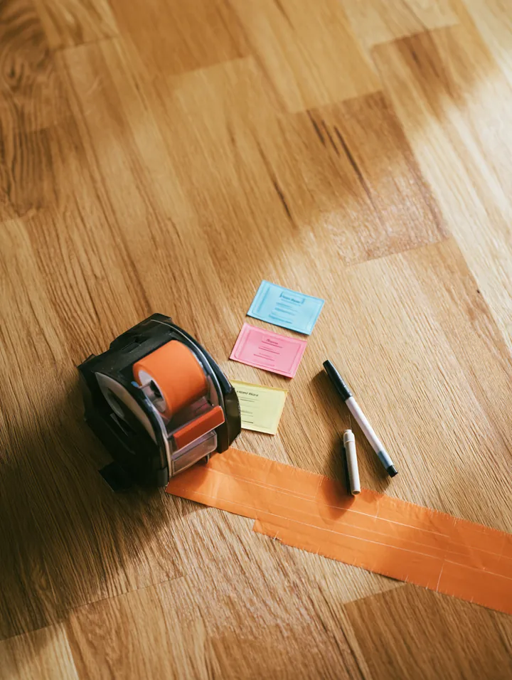 Top-down view of a packing tape dispenser, colorful room labels and a marker on the hardwood floor of an empty apartment
